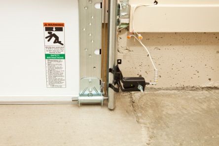Close-up of a properly installed garage door safety sensor with safety warning label and clean wiring on a concrete garage floor.
