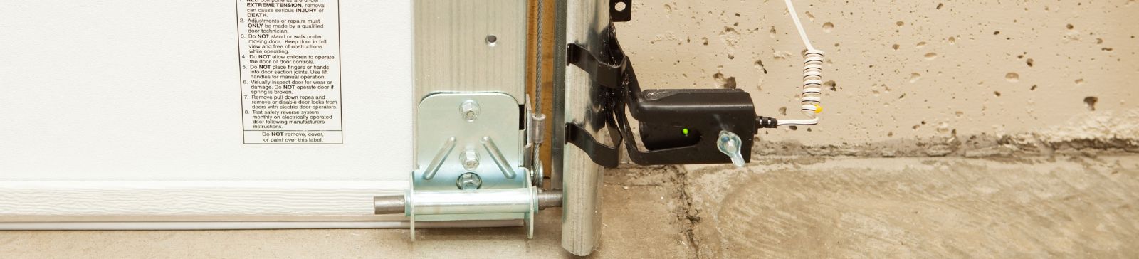 Close-up of a properly installed garage door safety sensor with safety warning label and clean wiring on a concrete garage floor.