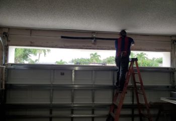 Technician on a ladder installing new black torsion springs above a white sectional garage door with palm trees visible outside in Winter Springs.