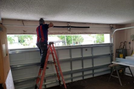Technician on a ladder installing new black torsion springs above a white sectional garage door with palm trees visible outside in Winter Springs.