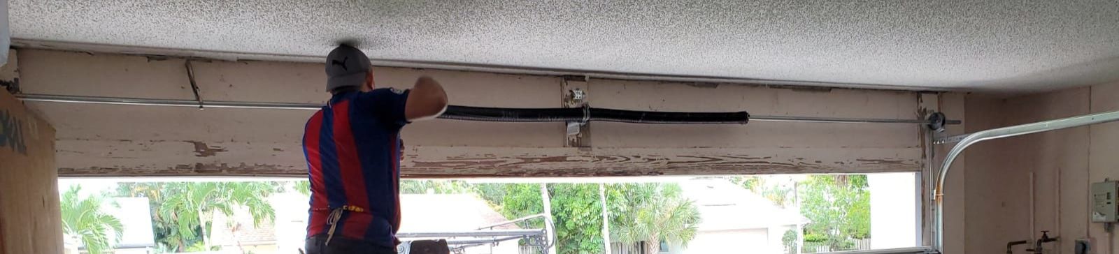 Technician on a ladder installing new black torsion springs above a white sectional garage door with palm trees visible outside in Winter Springs.